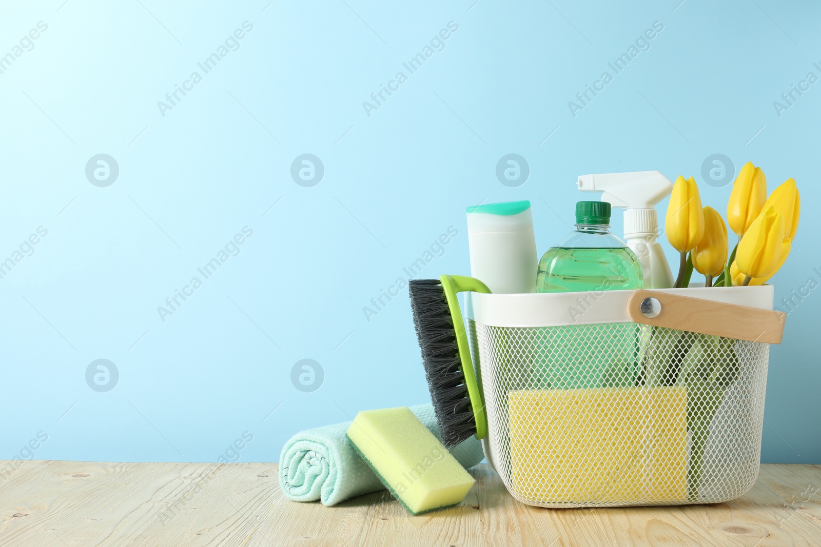 Photo of Basket with different cleaning supplies and beautiful spring flowers on wooden table against light blue background. Space for text