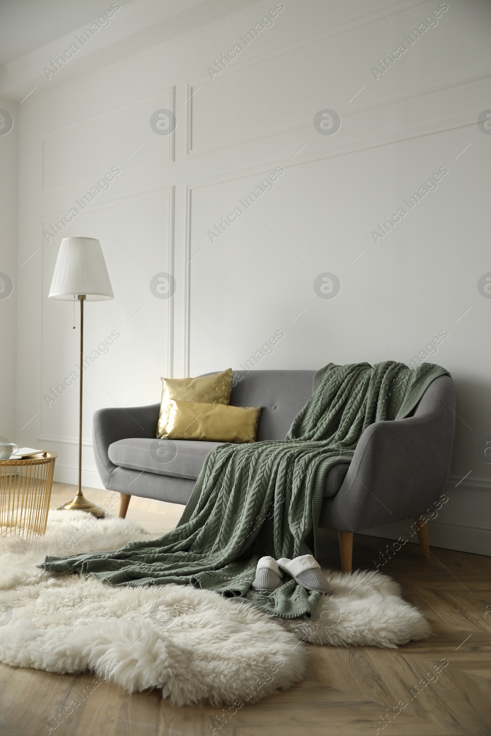 Photo of Stylish room interior with comfortable sofa, knitted blanket and lamp near white wall