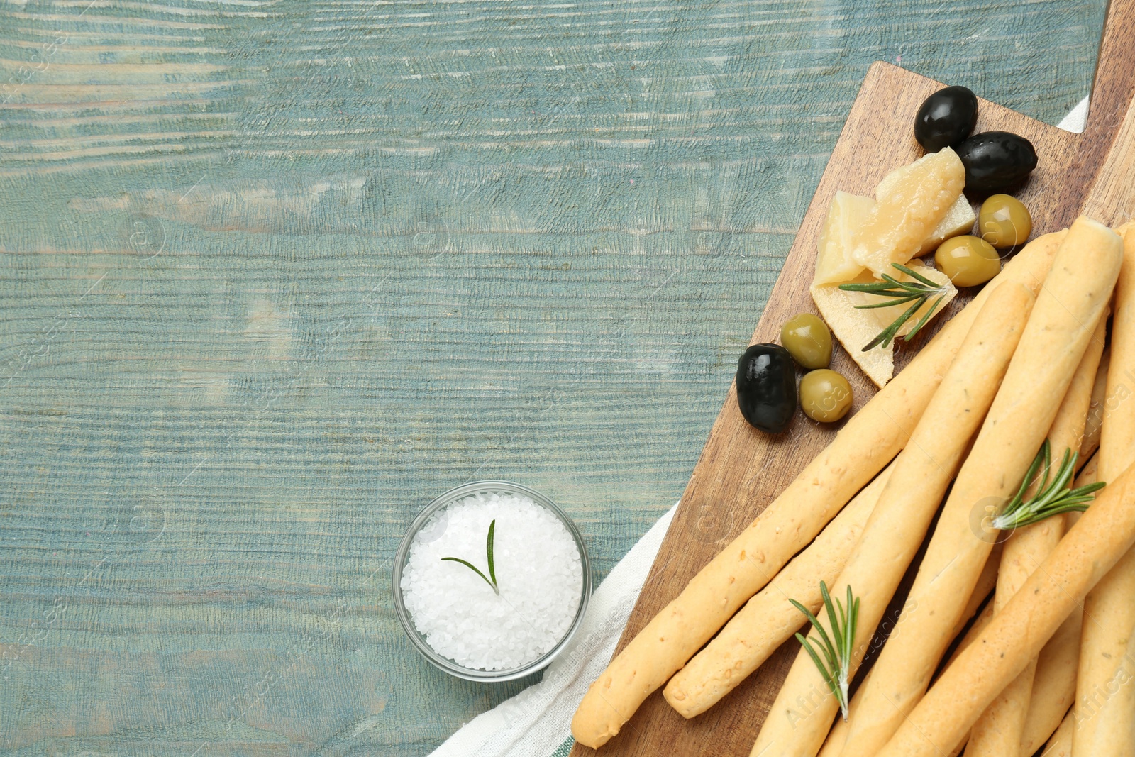 Photo of Tasty grissini with rosemary, cheese, olives and salt on light blue wooden table, flat lay. Space for text