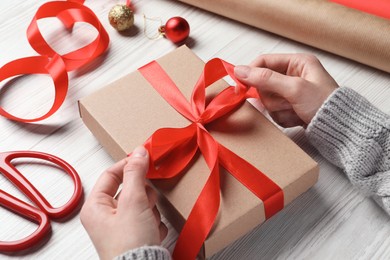 Christmas present. Woman tying ribbon bow on gift box at white wooden table, closeup