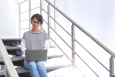 Young woman with modern laptop sitting on stairs indoors