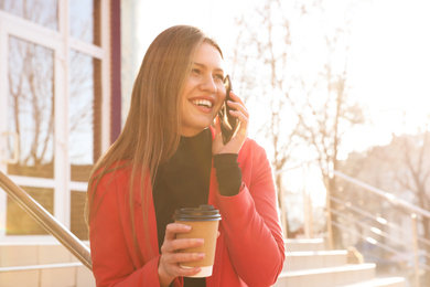 Photo of Businesswoman with cup of coffee talking on smartphone on city street in morning
