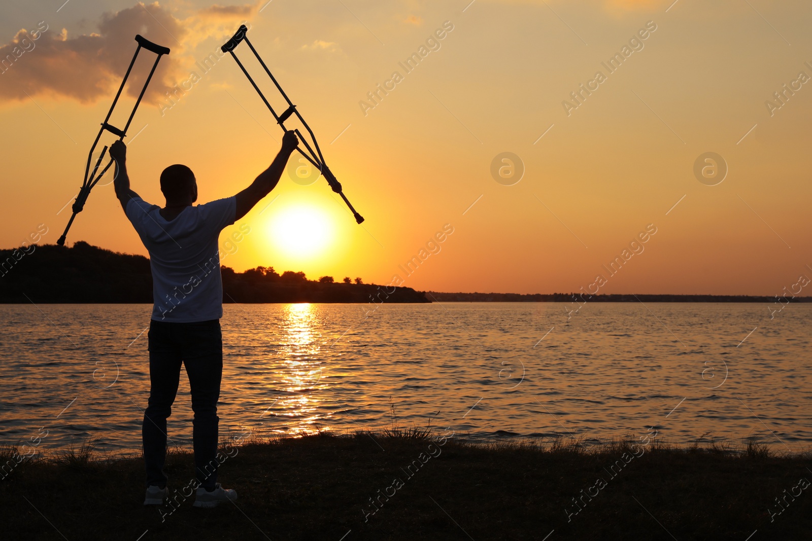 Photo of Man raising hands with underarm crutches up to sky near river at sunset, back view. Healing miracle