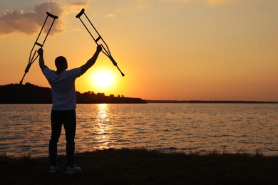 Photo of Man raising hands with underarm crutches up to sky near river at sunset, back view. Healing miracle