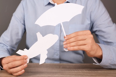 Photo of Insurance agent covering paper plane with umbrella cutout at table, closeup. Travel safety concept