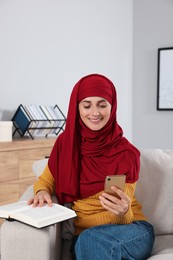 Muslim woman with book using smartphone on couch in room