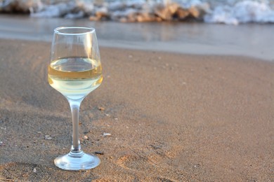 Glass of tasty wine on sand near sea, space for text