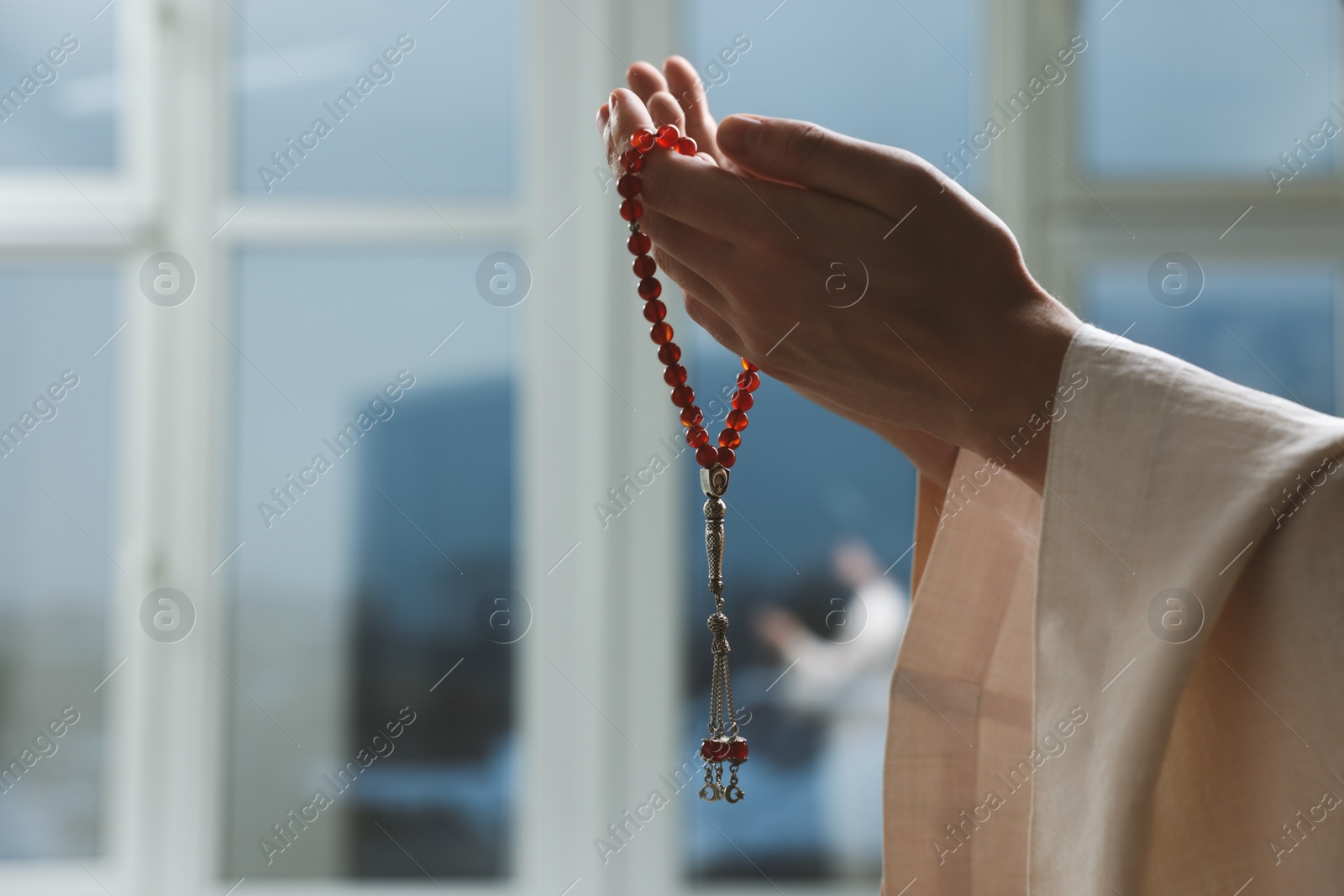 Photo of Muslim man with misbaha praying near window indoors, closeup. Space for text