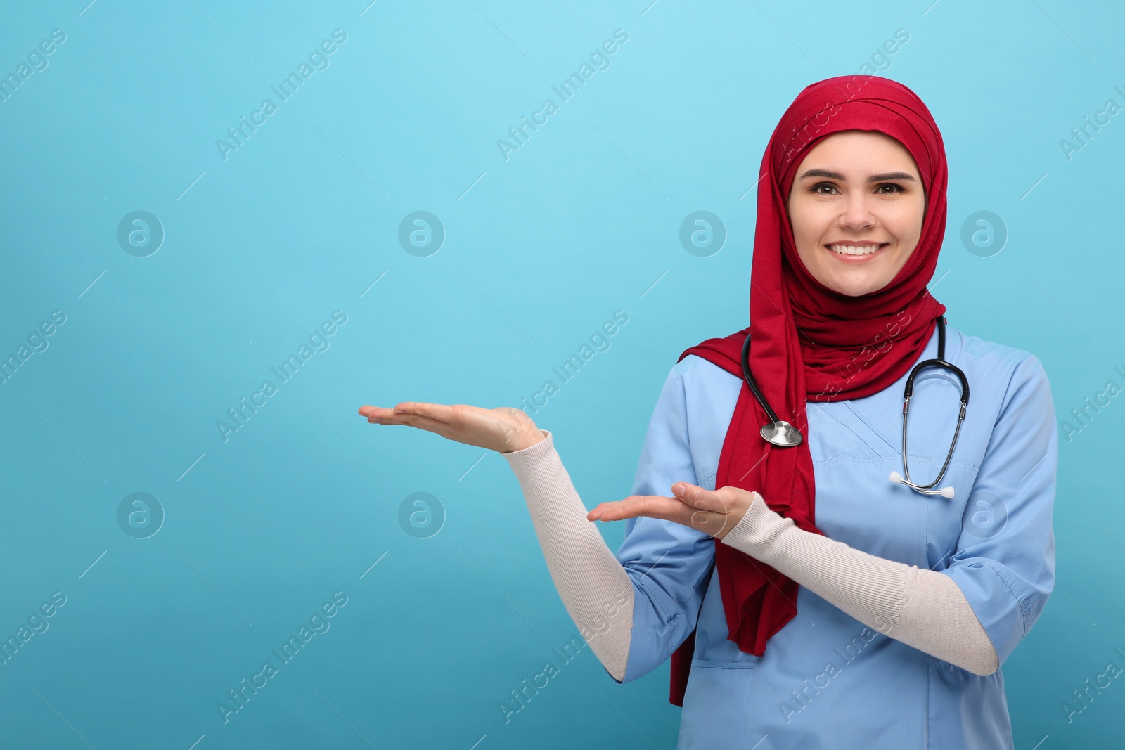 Photo of Muslim woman in hijab and medical uniform pointing at something on light blue background