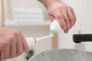 Photo of Man squeezing toothpaste from tube onto electric toothbrush indoors, closeup