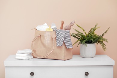 Photo of Mother's bag with baby's stuff and houseplant on white chest of drawers near beige wall