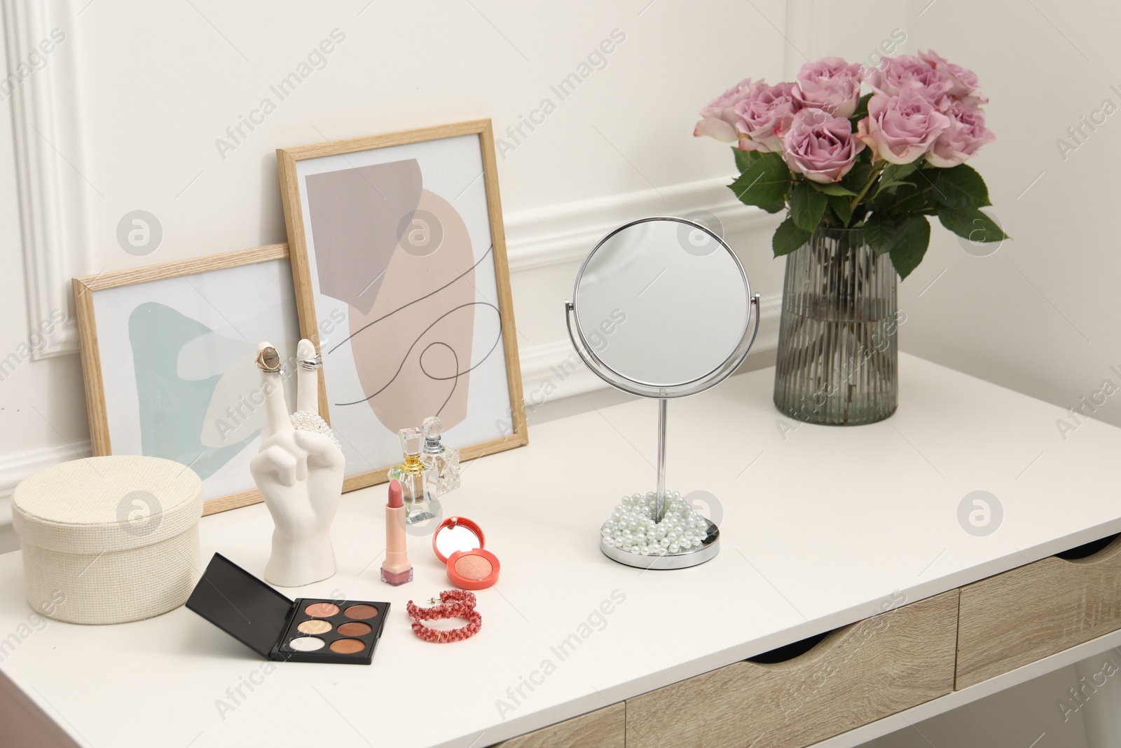 Photo of Mirror, cosmetic products, perfumes and vase with pink roses on white dressing table in makeup room