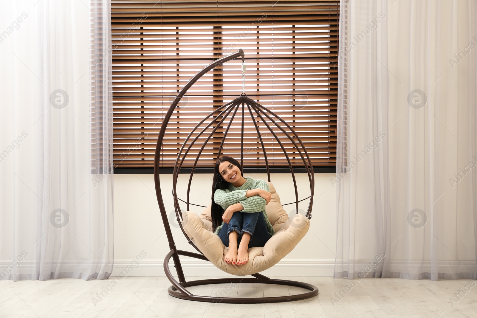 Photo of Young woman relaxing in hanging chair near window at home