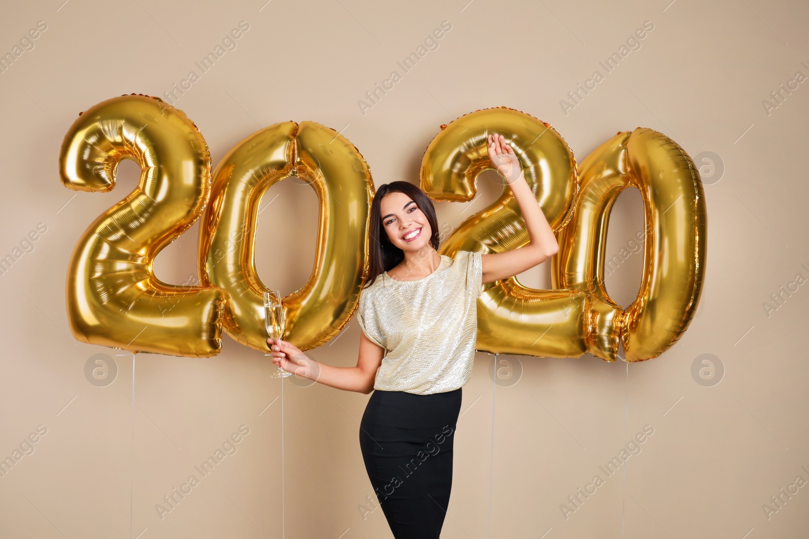 Photo of Happy young woman with glass of champagne near golden 2020 balloons on beige background. New Year celebration