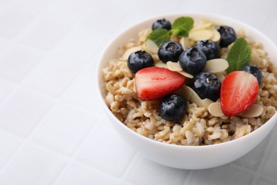 Photo of Tasty oatmeal with strawberries, blueberries and almond petals in bowl on white tiled table, closeup. Space for text