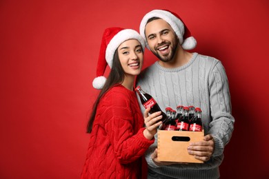 MYKOLAIV, UKRAINE - JANUARY 27, 2021: Young couple in Christmas hats holding crate with bottles of Coca-Cola on red background. Space for text