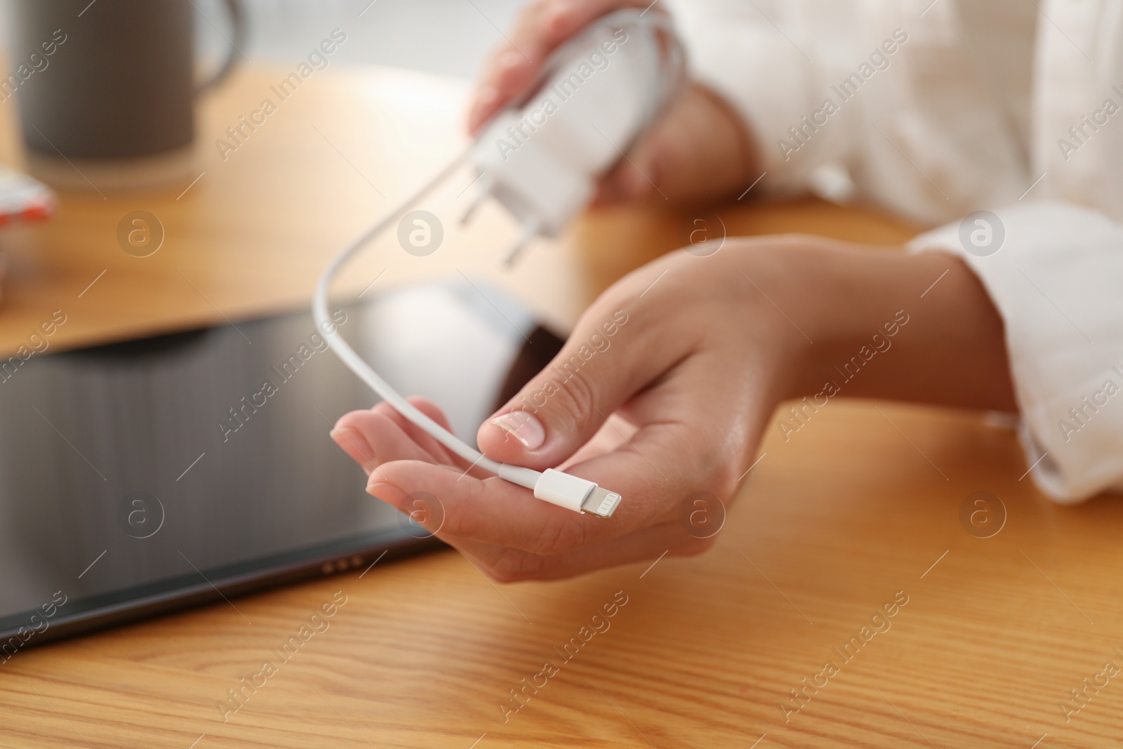 Photo of Woman holding charger near tablet at wooden table, closeup