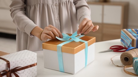 Photo of Woman decorating gift box with ribbon at white table indoors, closeup