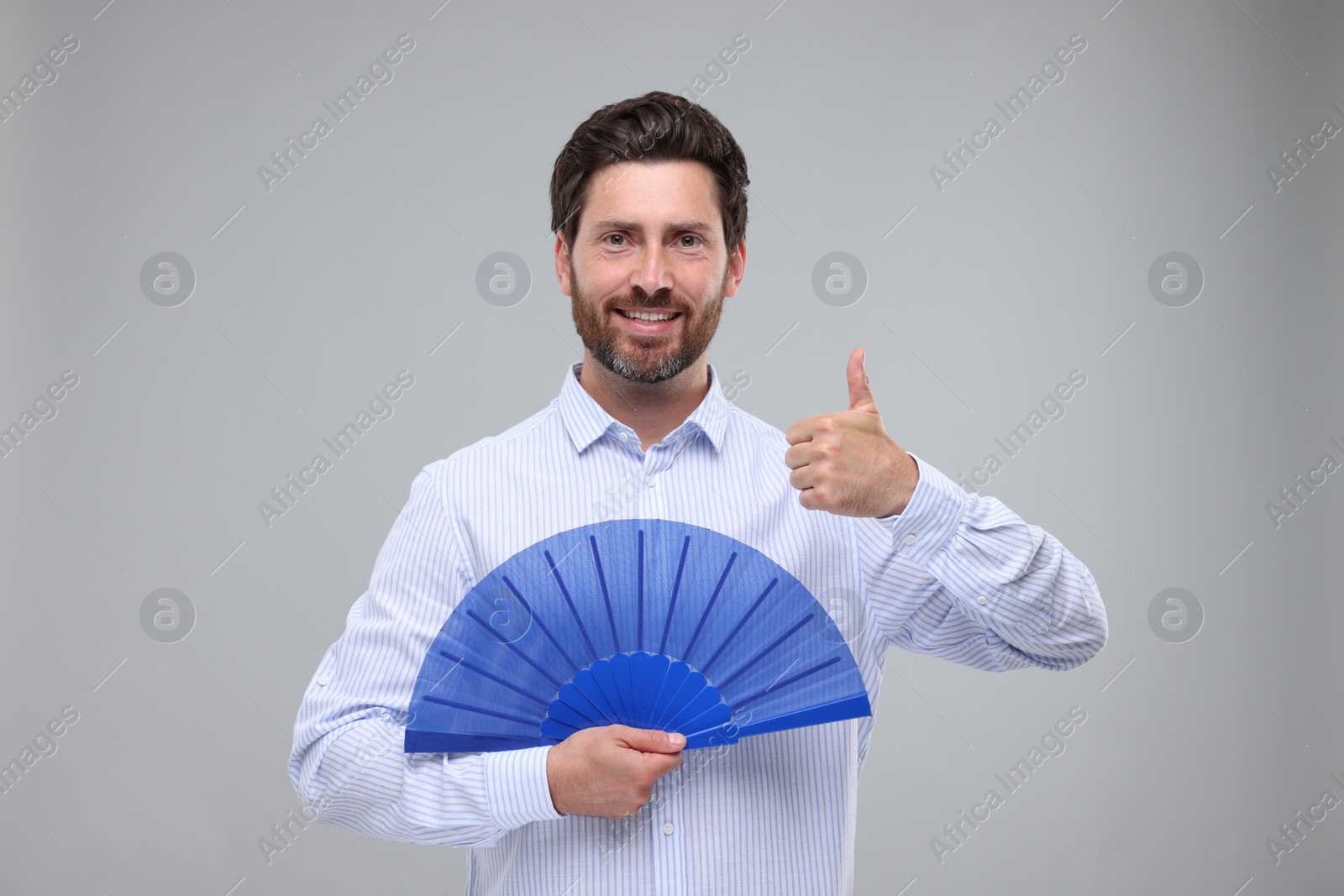 Photo of Happy man holding hand fan and showing thumb up on light grey background