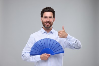 Happy man holding hand fan and showing thumb up on light grey background