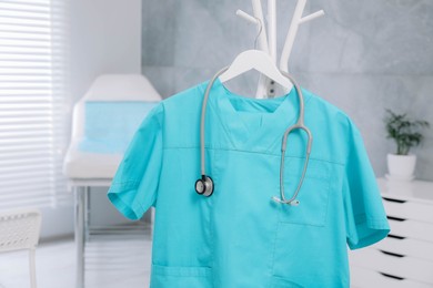 Turquoise medical uniform and stethoscope on rack in clinic, closeup