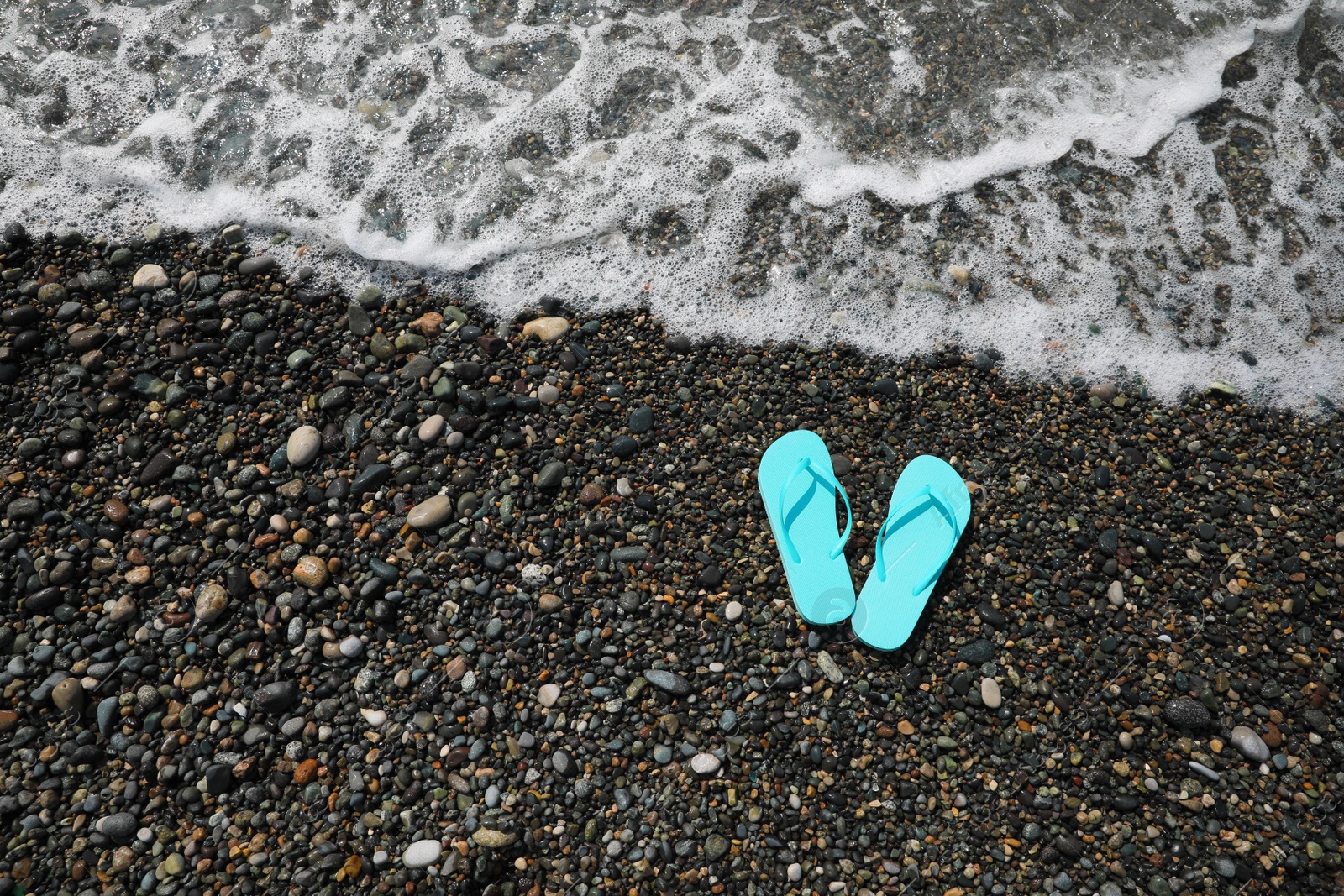 Photo of Stylish turquoise flip lops on pebble beach near beautiful sea wave, above view. Space for text