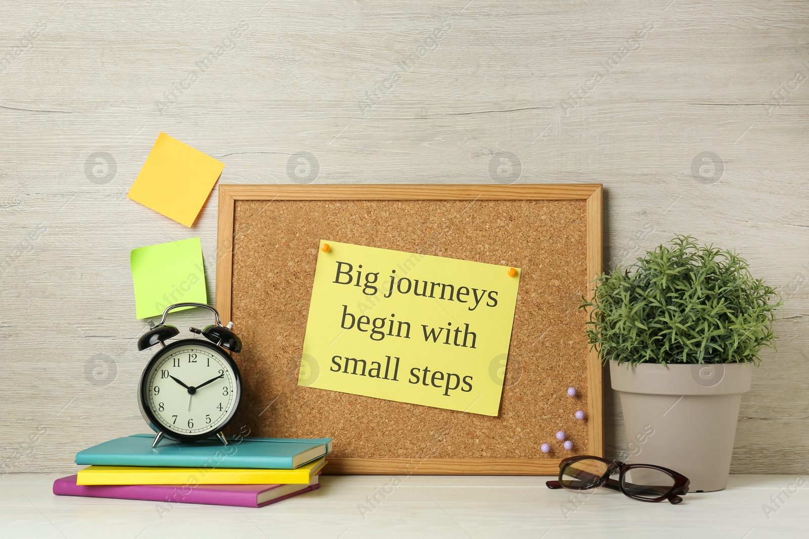 Photo of Cork board with motivational quote Big Journey Begin with Small Steps, notebooks, alarm clock and plant on white wooden table