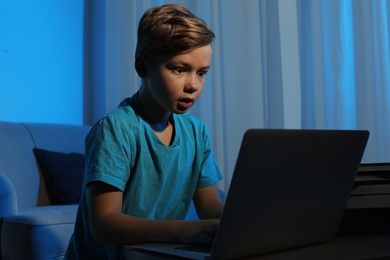 Photo of Shocked little child with laptop in dark room. Danger of internet