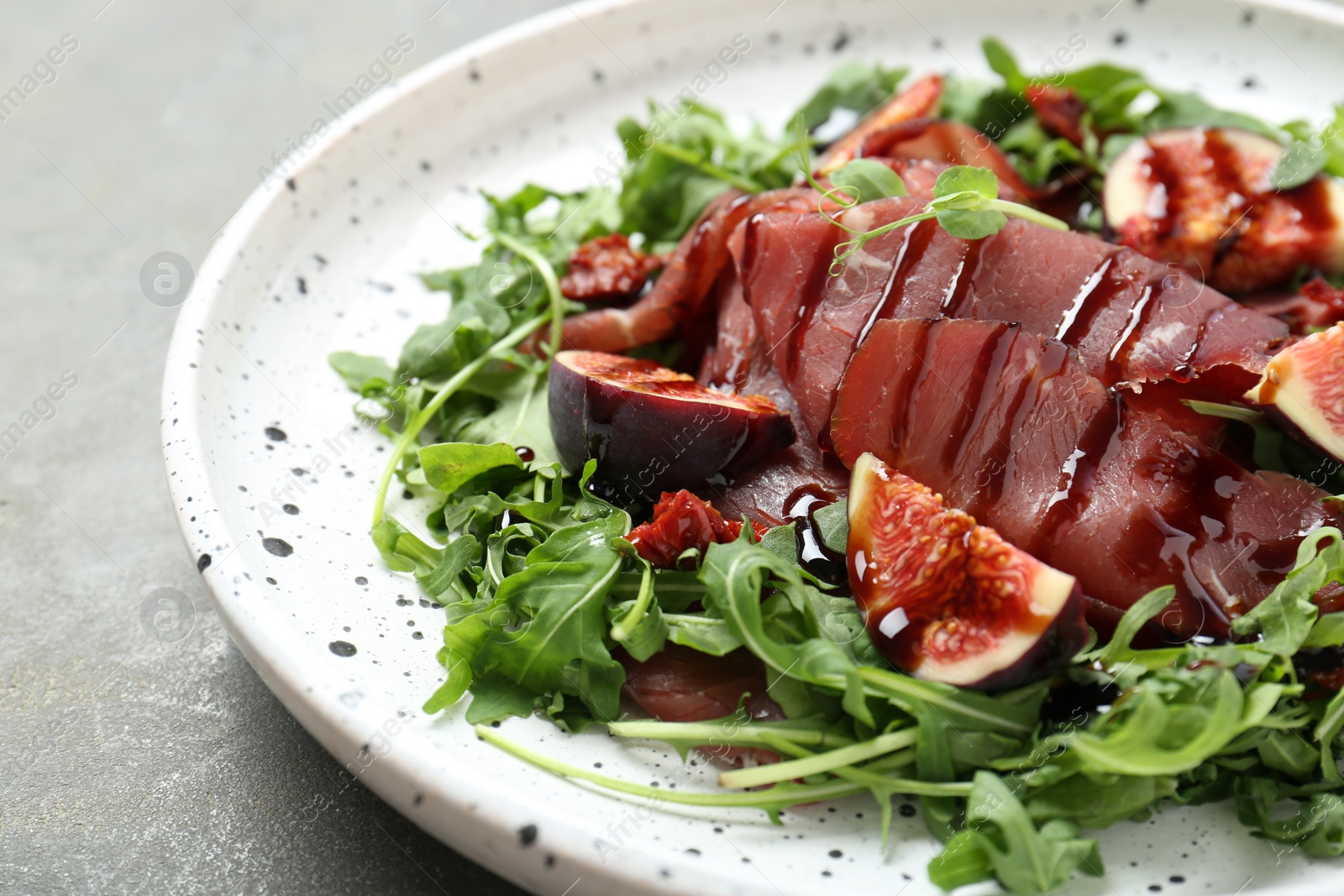 Photo of Plate of tasty bresaola salad with figs, sun-dried tomatoes and balsamic vinegar on grey table, closeup