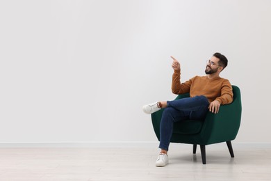 Handsome man pointing at something while sitting in armchair near white wall indoors, space for text