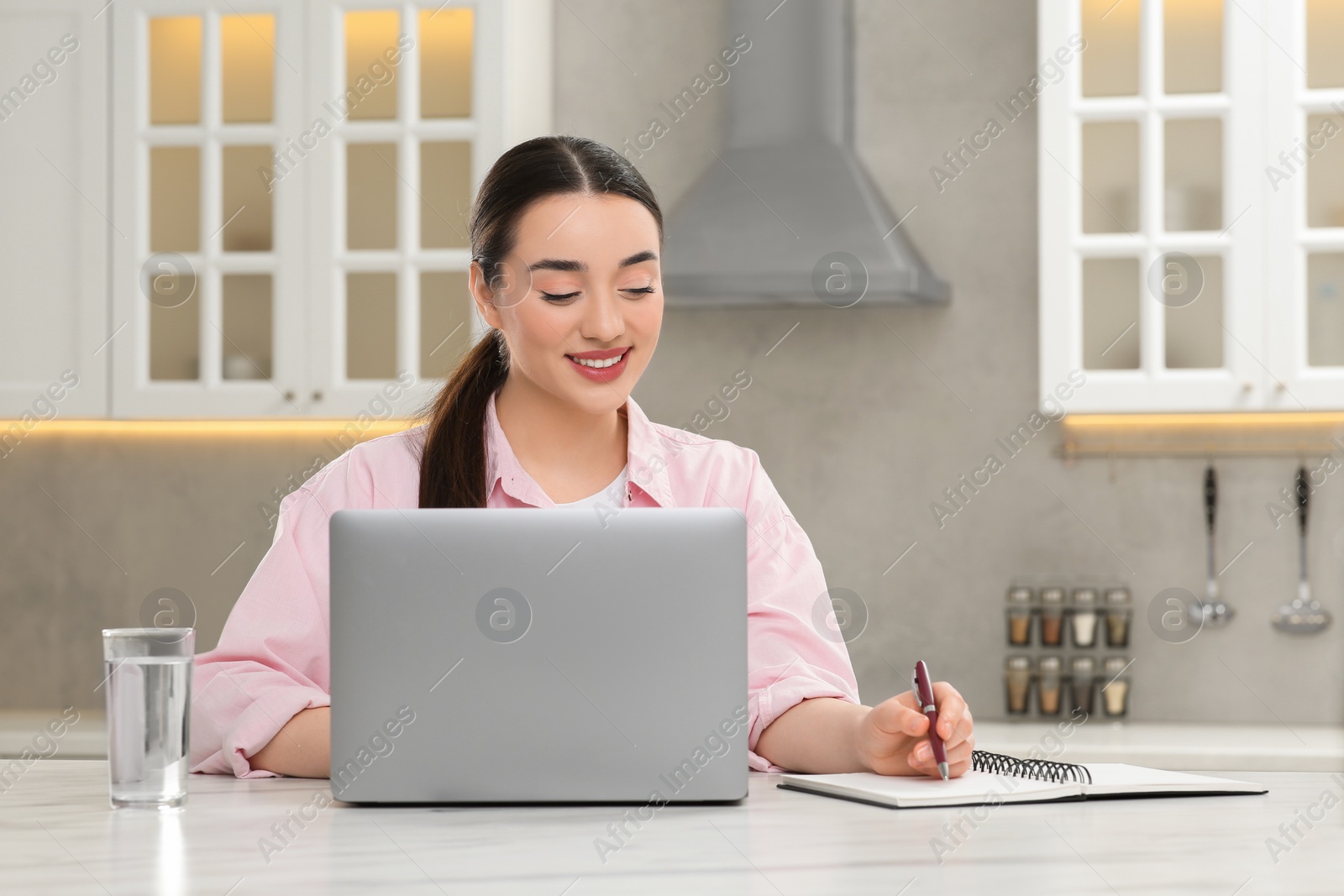 Photo of Woman writing something in notebook while using laptop at white table in kitchen