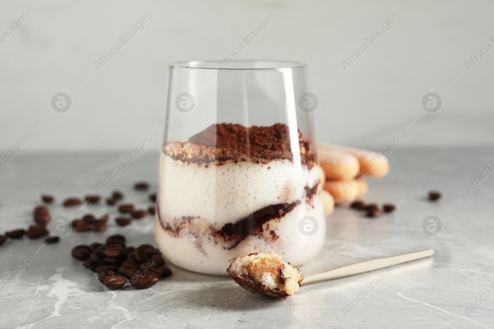 Photo of Tasty tiramisu in glass, coffee beans, spoon and biscuits on light grey table, closeup