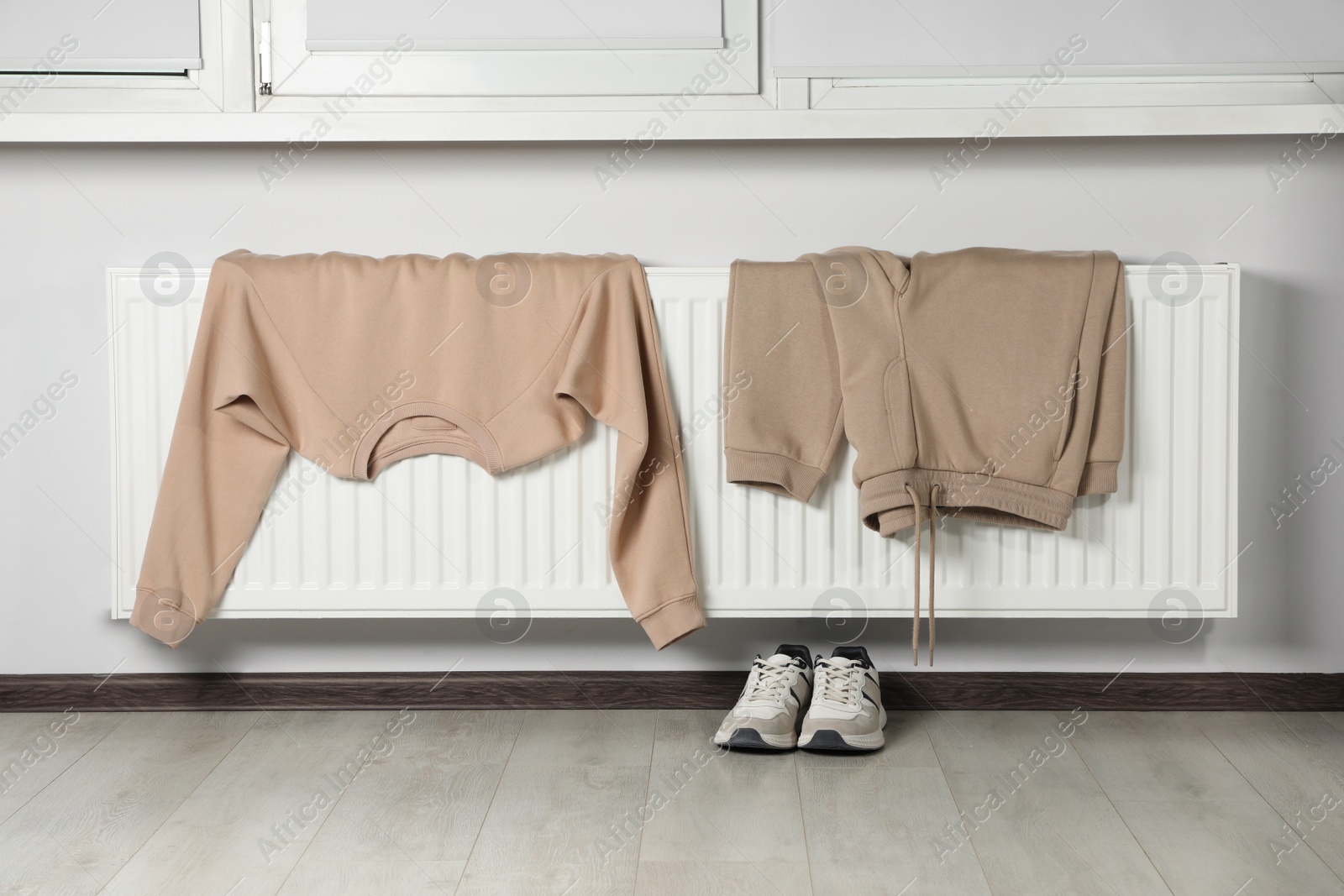 Photo of Heating radiator with sweatshirt, pants and sneakers near window indoors