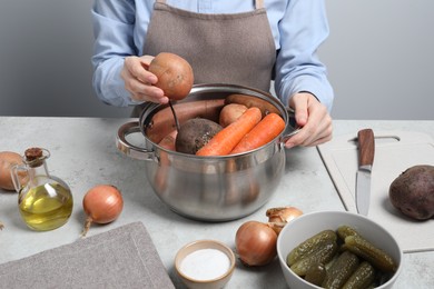 Photo of Woman putting potato into pot with fresh vegetables at white table, closeup. Cooking vinaigrette salad