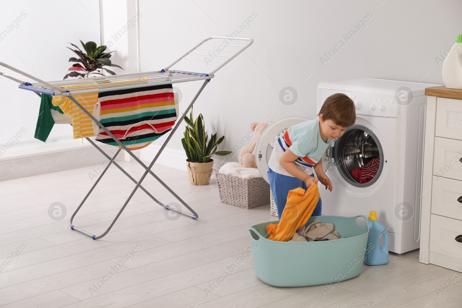 Photo of Cute little boy with laundry near washing machine and clothes drying rack indoors