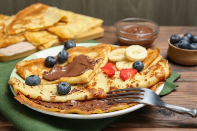 Photo of Tasty crepes with chocolate paste, banana and berries served on wooden table, closeup