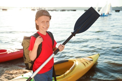 Photo of Happy boy with paddle near kayak on river shore. Summer camp activity