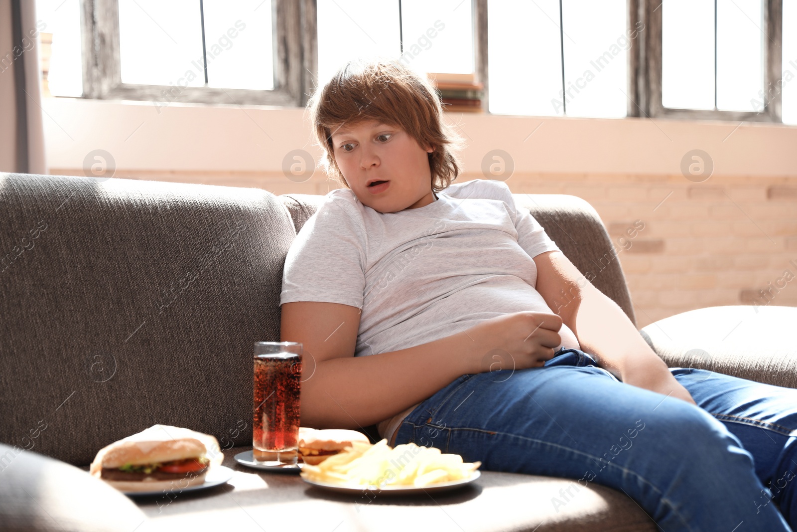 Photo of Emotional overweight boy with fast food on sofa at home