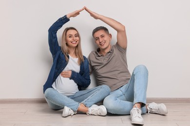 Photo of Young family housing concept. Pregnant woman with her husband forming roof with their hands while sitting on floor indoors