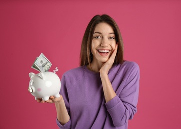 Happy young woman putting money into piggy bank on pink background