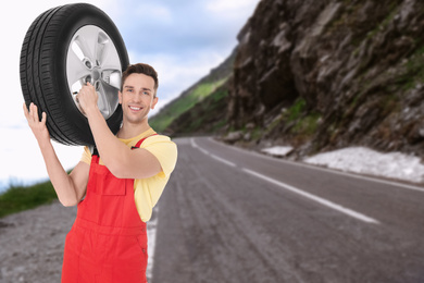 Mechanic in uniform holding car tire on road outdoors, space for text