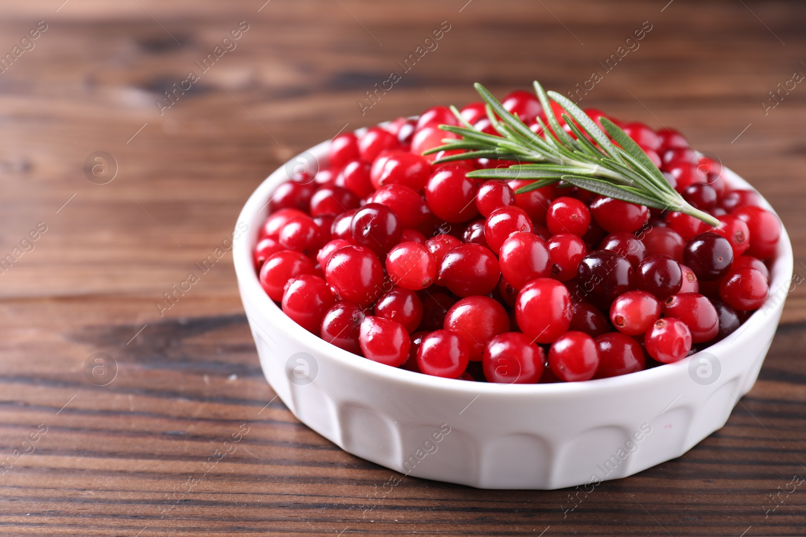 Photo of Fresh ripe cranberries and rosemary in bowl on wooden table, closeup