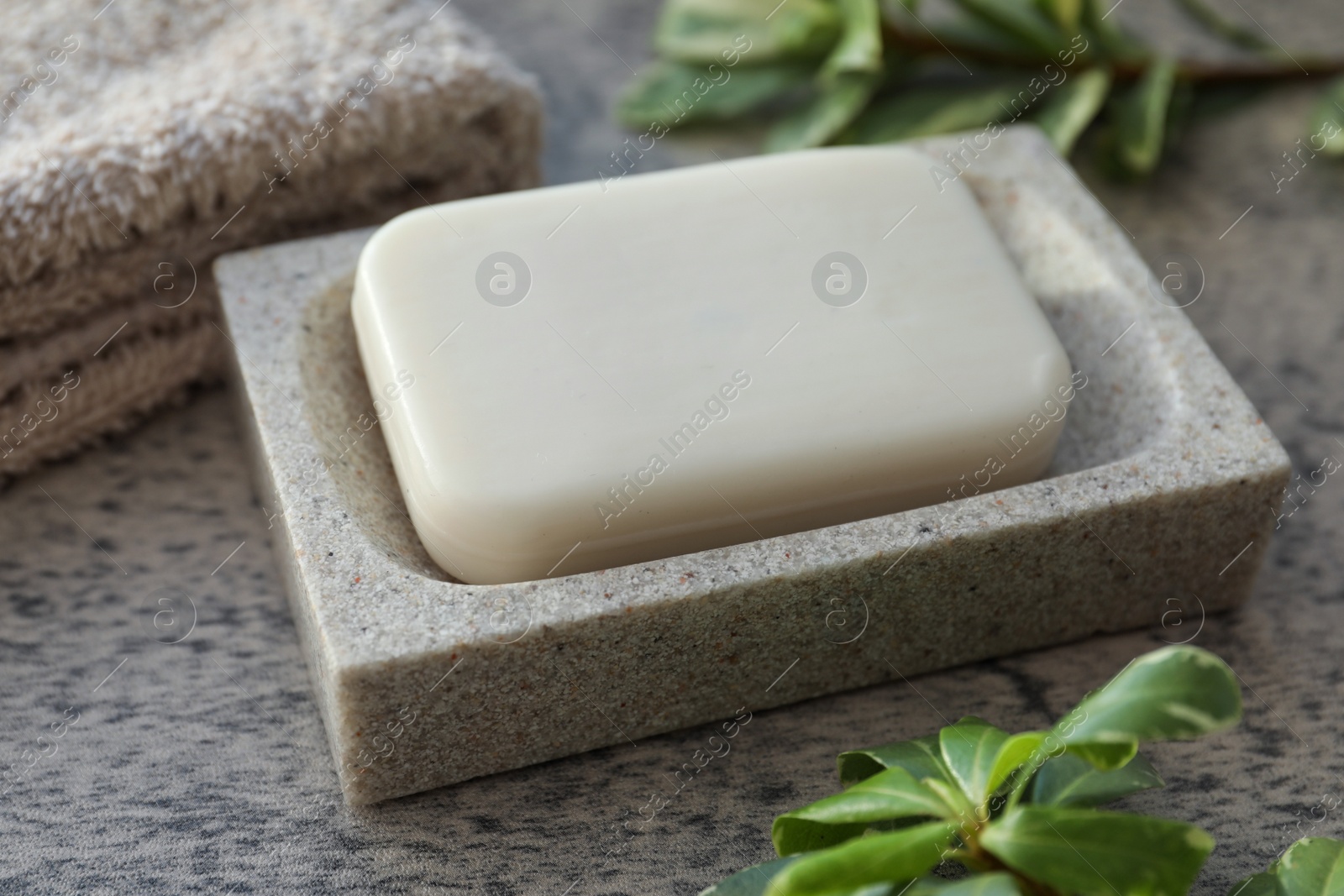 Photo of Dish with soap bar, terry towel and green plants on light grey textured table, closeup