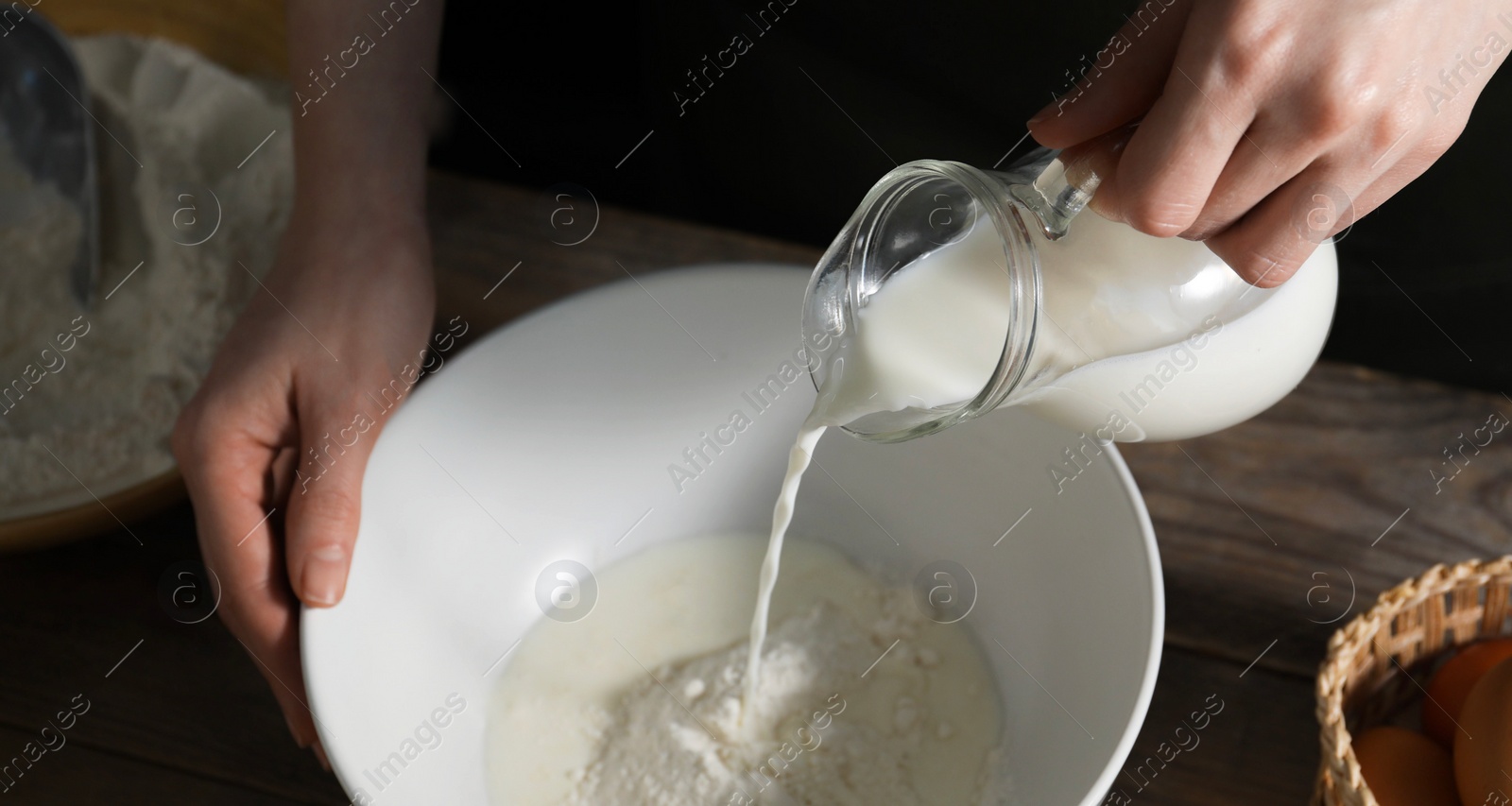 Photo of Making bread. Woman pouring milk into bowl at wooden table on dark background, closeup