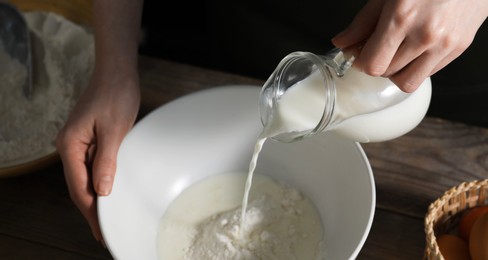 Making bread. Woman pouring milk into bowl at wooden table on dark background, closeup