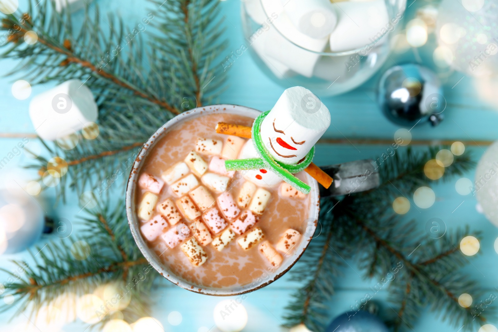 Image of Funny marshmallow snowman in cup of hot drink on light blue wooden table, top view. Bokeh effect 
