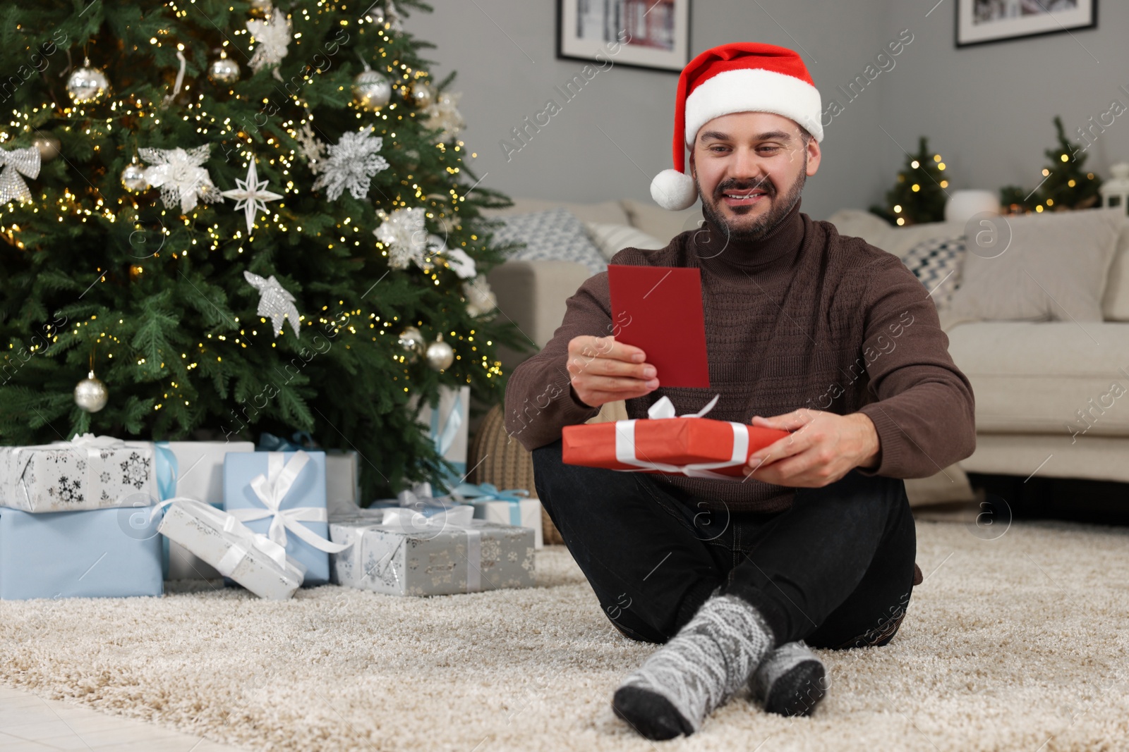 Photo of Happy man in Santa hat with Christmas gift reading greeting card at home