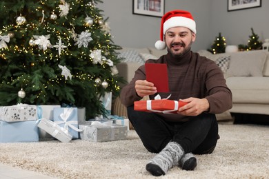 Happy man in Santa hat with Christmas gift reading greeting card at home
