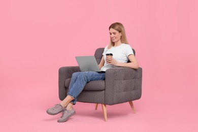 Happy woman with laptop and cup of drink sitting in armchair against pink background