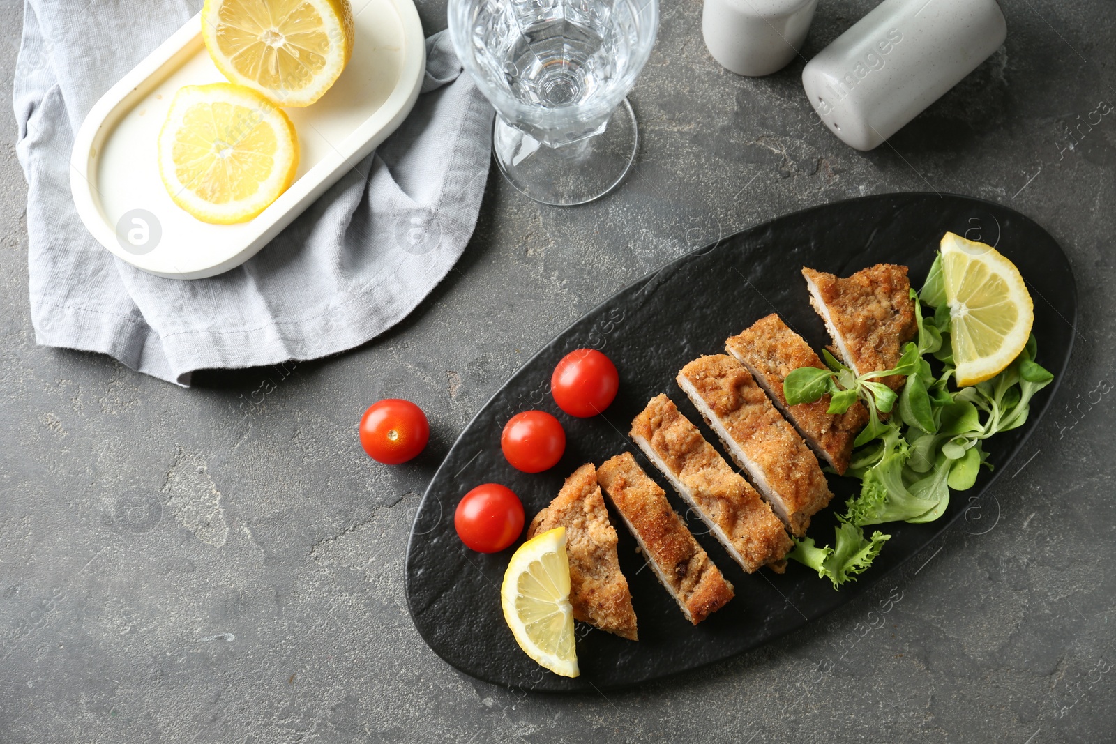 Photo of Tasty cut schnitzel served with tomatoes, greens and lemon on grey textured table, flat lay